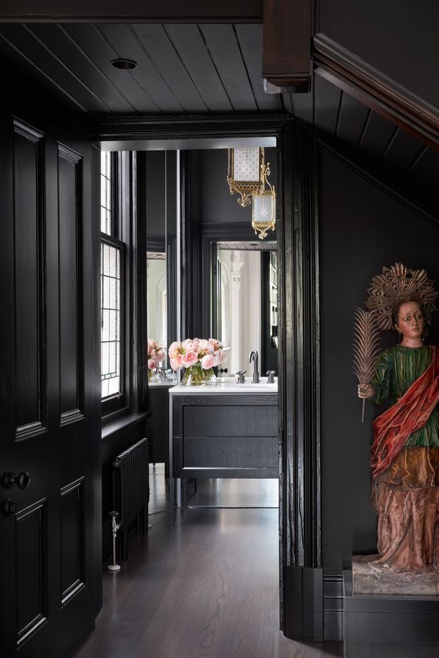 A dramatic, narrow interior view through a black-painted paneled doorway into a compact bathroom. The space features black wall panelling and ceiling boards, dark timber flooring, a floating dark vanity with a white basin, a large mirror, tall divided-light windows, and an ornate gold pendant light. A colourful sculptural artwork occupies a built-in niche on the right, while pink flowers add a small soft accent. The overall composition is moody, richly detailed, and historic in character with contemporary bathroom fixtures.