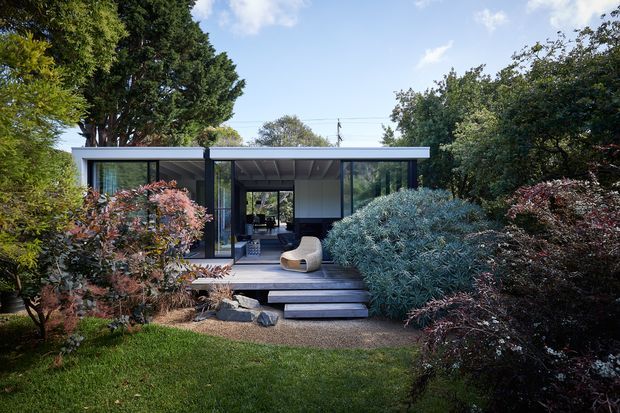 A low, modern pavilion-style house set within a mature landscaped garden. The facade features extensive dark-framed glazing, a flat roof with a projecting eave, and a central covered entry reached by broad dark timber steps. Dense shrubs and established trees frame the building, while a single sculptural outdoor chair is visible beneath the sheltered entry.