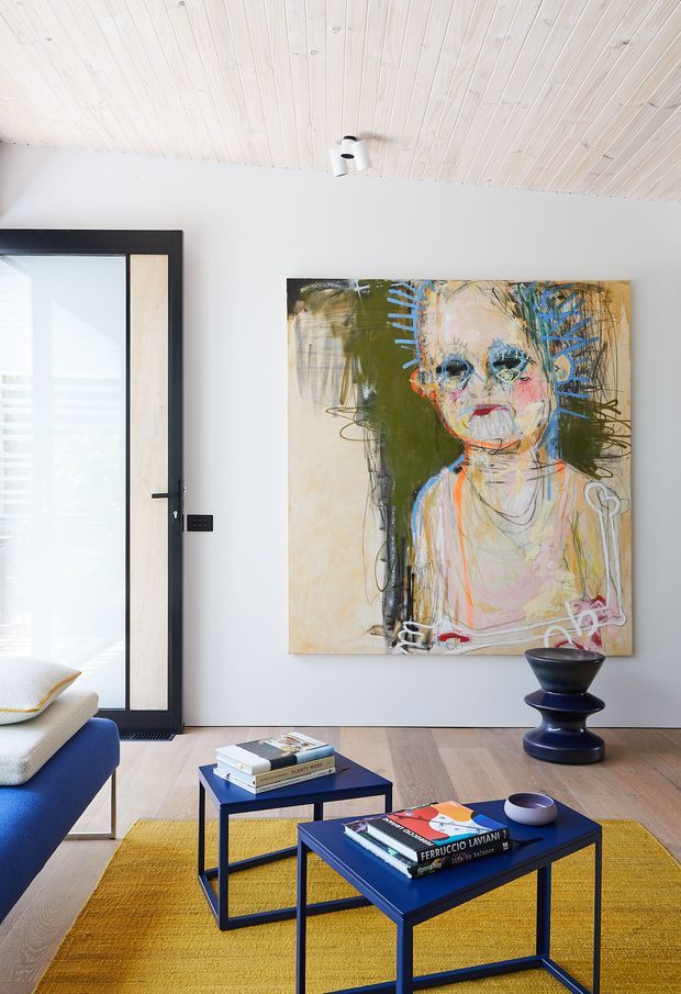 Bright contemporary living room with a pale timber-lined ceiling, white walls, light timber flooring, and a black-framed glazed door. A large expressive abstract portrait dominates the feature wall, while blue square coffee tables, a mustard rug, a sculptural dark side table, and part of a blue sofa create a bold, curated interior.