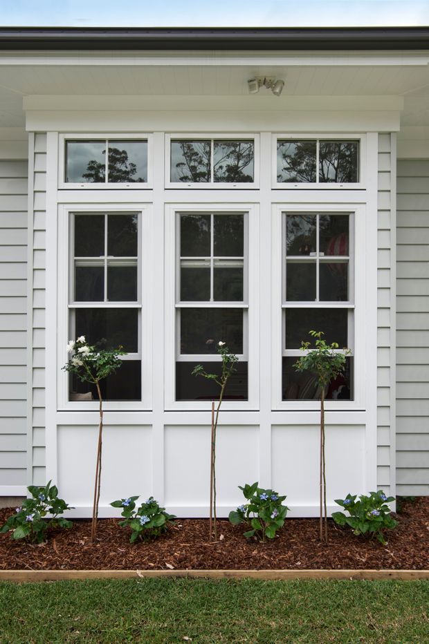 Front-facing exterior detail of a traditional white-painted house facade, featuring three tall double-hung windows with decorative upper transoms, white trim, and light grey horizontal weatherboard cladding. A covered soffit with a small exterior spotlight projects above the windows. Mulched garden beds with small shrubs and flowering plants run along the base.