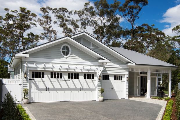 Exterior view of a light-painted, gable-roofed residential building dominated by a large multi-door garage, with a covered entry or porch to the right. The facade features horizontal weatherboard cladding, white trim, decorative garage door windows, and a circular upper gable window. A paved driveway and mature trees surround the property.