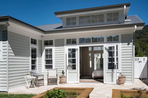 Exterior view of a light grey weatherboard home with white trim, a broad gabled roof, and a raised upper-level dormer. The open French doors reveal a bedroom, while the paved terrace includes outdoor seating and planters, with a small garden bed in the foreground.