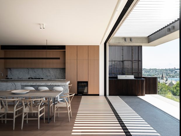 A bright, open-plan contemporary kitchen and dining area opening onto a covered outdoor terrace. The interior features warm timber cabinetry, a dark stone or marble-look splashback and island, a long dining table with light timber chairs, and extensive floor-to-ceiling glazing. Strong linear shadows from the external louvred roof and screen fall across the timber floor and terrace. A distant urban hillside view is visible beyond the outdoor entertaining area.