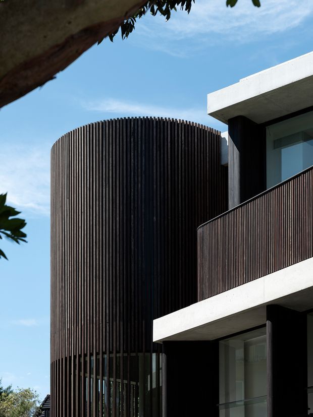 A close-up architectural exterior view of a contemporary multi-storey building. The facade combines a rounded volume wrapped in closely spaced dark vertical slats with white concrete slab edges, dark-framed glazing, and a projecting balcony with matching vertical balustrade screening. The composition emphasizes strong geometric forms, vertical rhythm, exposed concrete, and contrast against a clear blue sky, with tree branches framing the view.