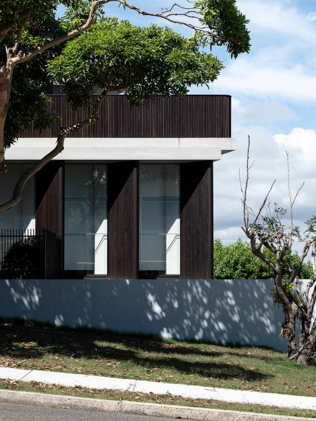 Exterior view of a contemporary multi-storey residence, featuring a dark vertical timber-clad upper facade, deep projecting concrete slab, tall glazed openings with angled dark fins, and a pale rendered boundary wall. Mature trees and landscaped lawn frame the street-facing elevation.