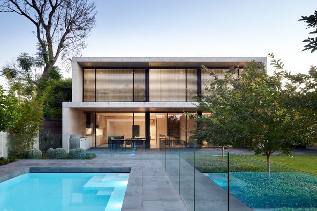 Contemporary two-storey residence viewed from the rear garden, with a rectilinear concrete-toned facade, extensive glazing screened by horizontal blinds, a covered outdoor dining area, paved terrace, lawn and a swimming pool in the foreground.
