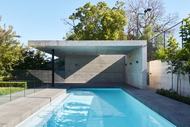 Modern residential outdoor pool area with a rectangular turquoise swimming pool enclosed by pale concrete walls and a projecting flat-roofed concrete pavilion. Dark slender structural posts, frameless glass pool fencing, large-format paving, clipped hedging and mature trees create a clean architectural landscape setting. Strong sunlight casts geometric shadows across the concrete wall and pool deck.