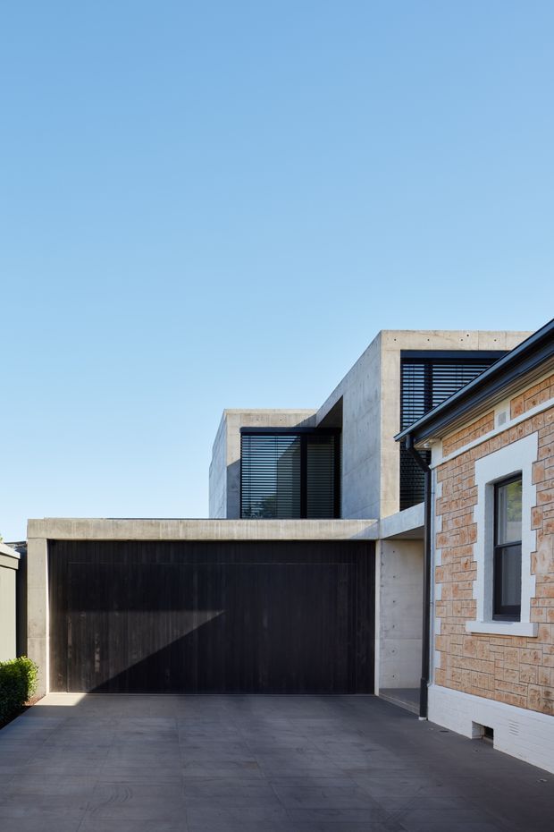 Exterior view of a contemporary multi-level residential addition, showing a large dark timber garage door, broad concrete-paved driveway, exposed concrete box forms, black louvred glazing, and an adjoining older brick structure with a pitched roof. The composition highlights the contrast between the modern concrete addition and the existing masonry building under a clear blue sky.