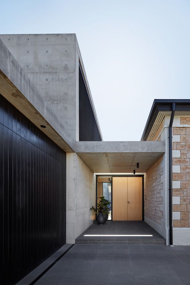 Exterior view of a contemporary residential entryway, featuring a recessed timber front door beneath a raw concrete canopy. The approach is paved in large dark grey tiles, with a black vertically ribbed garage door on the left and pale masonry cladding on the right. Strong geometric forms, concealed lighting, and contrasting concrete, dark metal, timber, and stone create a refined modern facade.