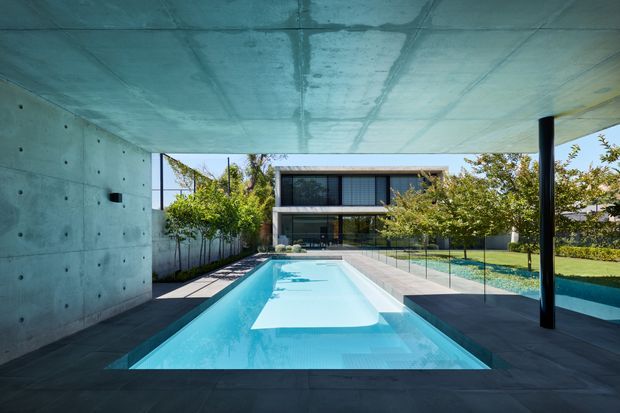 A modern residential courtyard viewed from beneath a deep cantilevered concrete roof or terrace. A long rectangular swimming pool extends toward a two-storey glazed house, with dark tiled paving, a raw concrete wall and ceiling, slender black support columns, glass pool fencing, and landscaped garden areas.