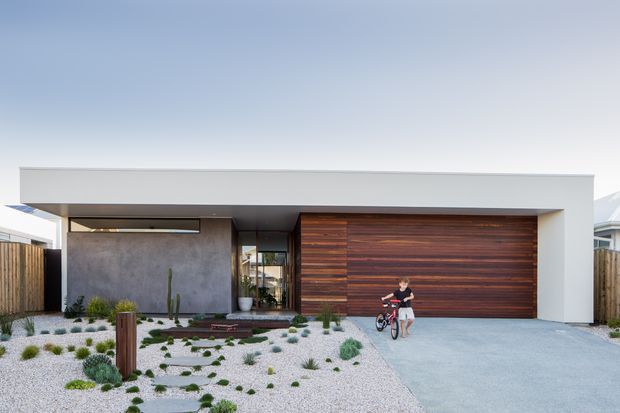 Front exterior view of a contemporary single-storey residence with a broad flat roof canopy, white rendered facade, dark timber garage door, concrete driveway, and a low-maintenance gravel garden with drought-tolerant planting. A child with a bicycle is present near the garage.