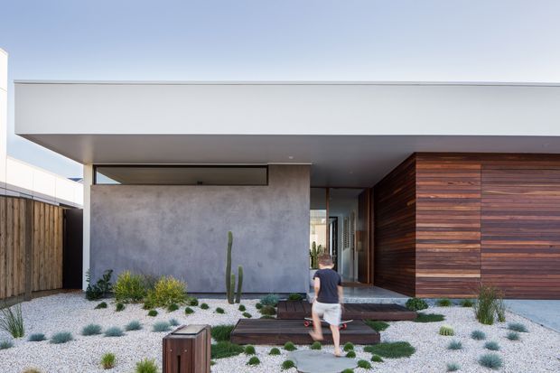 A contemporary single-storey residential facade with a broad flat roof and deep overhang. The entry is framed by smooth grey concrete-rendered walls and warm horizontal timber cladding, with a recessed glazed doorway and timber stepping platforms leading through a minimalist drought-tolerant gravel landscape. The composition is clean and rectilinear, with strong indoor-outdoor connections and understated detailing.
