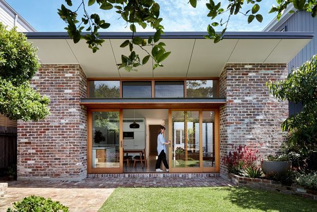 Exterior view of a contemporary single-storey residential extension or courtyard-facing home. The facade combines exposed red brick walls, extensive timber-framed glazing and doors, and a broad low-pitched flat roof with a sheltered overhang. A landscaped garden and paved terrace sit in front, while the interior is visible through the glazing.