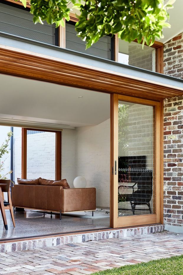 A bright architectural view of a brick-clad courtyard or patio looking into a contemporary living room. Large timber-framed glazed openings create a strong indoor–outdoor connection, with a warm brown leather sofa, white-painted brick interior walls, polished concrete or terrazzo-like flooring, and exposed timber detailing. A paved brick terrace and leafy planting frame the foreground.