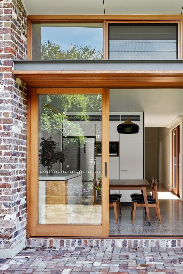 View from an exterior paved courtyard through a large timber-framed glazed opening into a bright dining and kitchen area. The facade combines exposed red brick with warm timber window and door frames, while the interior features a timber dining table and chairs, white built-in kitchen cabinetry, a black pendant light, and polished aggregate-style flooring. Reflections reveal greenery and additional glazing, and the upper-level opening includes timber framing and a horizontal louvre or blind.