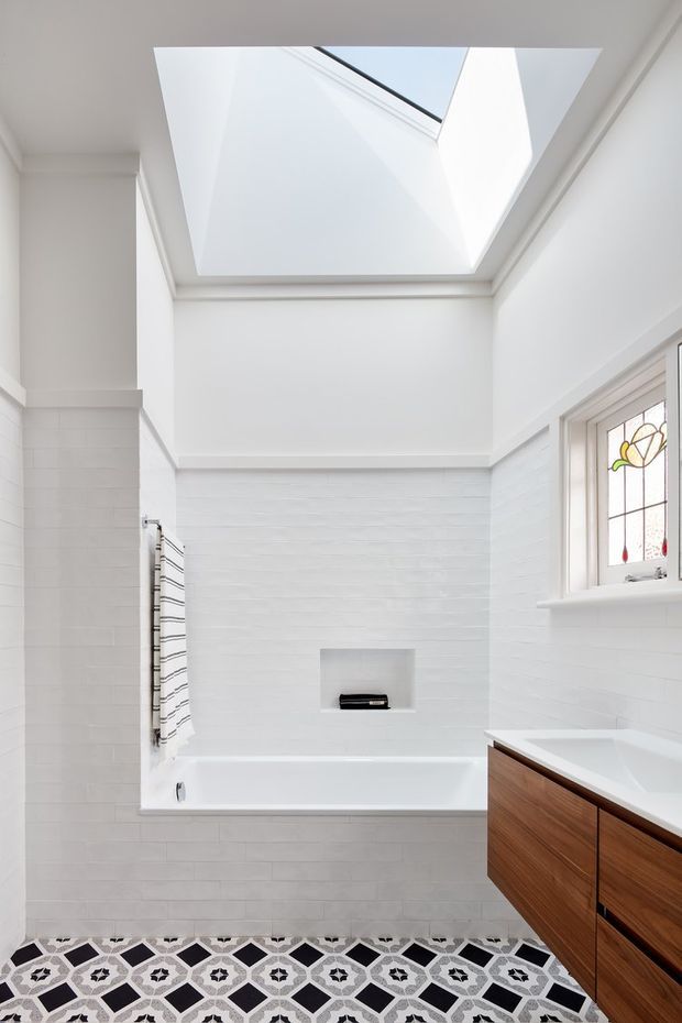 Bright white bathroom with a built-in bathtub beneath a large skylight, white tiled walls, patterned geometric floor tiles, a wall-mounted towel warmer, stained-glass window, and a floating timber vanity with a white benchtop. The room has abundant natural light and clean, refined detailing.