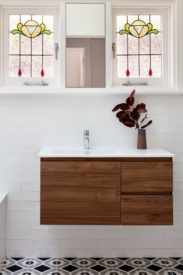 A bright, refined bathroom vanity area with a floating dark timber wall-mounted cabinet, white countertop and integrated basin, chrome tapware, and white horizontal wall tiles. Two decorative stained-glass windows with floral motifs flank a central mirror above the vanity. The patterned geometric floor tiles add a distinctive decorative accent.