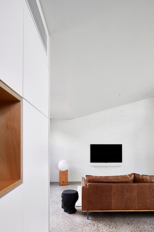 A minimalist living room viewed from an adjacent area, featuring a brown leather sofa facing a wall-mounted black television on a white painted brick wall. The room has a pale terrazzo floor, a white ceiling with recessed downlights, and integrated white wall cabinetry with a warm timber-lined niche at left. A small sculptural black side table and a timber pedestal with a round white lamp add restrained accents.