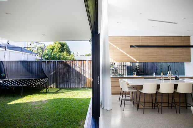 Bright contemporary open-plan kitchen and dining area opening directly onto a compact grassed backyard through wide floor-to-ceiling glazing and sliding doors. The kitchen features a long white island with integrated sink and seating, pale cabinetry, a timber-look wall panel, dark linear rangehood, and a polished light terrazzo-style floor. A trampoline and timber boundary fence are visible outdoors.