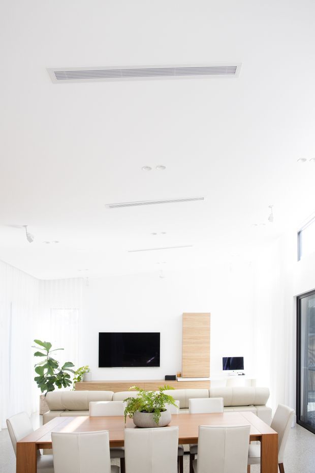 Bright, minimalist open-plan living and dining area with white walls and ceiling, sheer curtains, light timber built-in media cabinetry, a wall-mounted television, cream upholstered seating, and a large timber dining table with white chairs. Recessed and linear air-conditioning vents contribute to the clean contemporary fit-out, while large glazing on the right brings in abundant daylight.