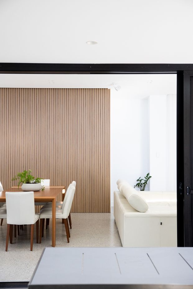Bright open-plan interior viewed through a large black-framed opening, with a dining area and timber dining table on the left, a white upholstered sofa on the right, a full-height vertical timber slat feature wall, white walls and ceiling, and a pale terrazzo-like floor. Minimal contemporary furnishings and indoor plants create a clean, warm-neutral setting.