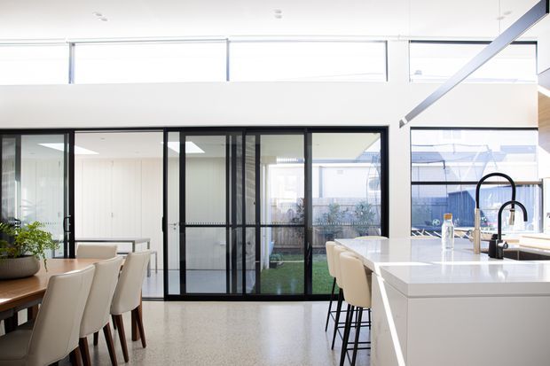 Bright contemporary open-plan kitchen and dining area with a large white island, black gooseneck tap, white upholstered bar stools, and a timber dining table with matching chairs. Black-framed glazed sliding doors open toward a landscaped courtyard, while high-level windows and a partially vaulted ceiling bring in abundant natural light. The floor appears to be a polished light terrazzo or concrete-like finish.