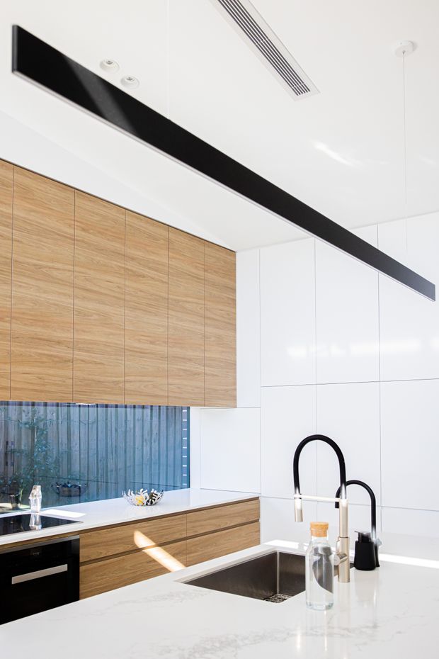Bright contemporary kitchen featuring white full-height wall panels, warm timber-grain cabinetry, a white stone island benchtop with an inset stainless-steel sink, matte black arched tapware, black appliances, and a prominent linear black ceiling light. A large window provides natural light and views to greenery outside.