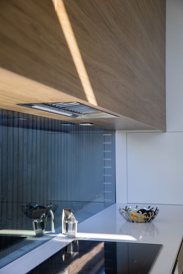 Close-up of a sleek contemporary kitchen featuring a dark timber-look overhead cabinet or ceiling panel, integrated stainless-steel rangehood, glossy white cabinetry and benchtop, black glass induction cooktop, and a reflective dark glass splashback. Strong natural light creates diagonal shadows across the cabinetry and work surface.