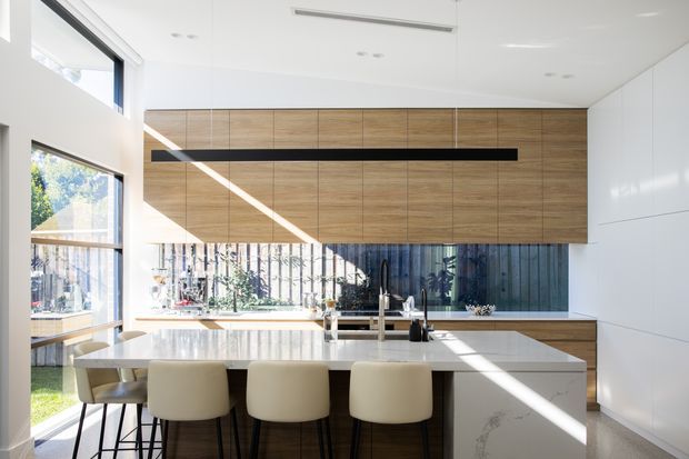 Bright contemporary kitchen with a large white island and integrated seating for four, extensive warm timber overhead cabinetry, white built-in cabinetry, and a dark reflective splashback. Full-height glazing and clerestory-style windows bring in strong natural light, with a long linear pendant above the island.