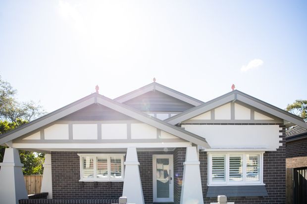 Front exterior view of a multi-gabled suburban home with dark brick walls, white rendered trim, timber-style gable detailing, decorative finials, and white shuttered windows. The façade is brightly sunlit and presents a traditional cottage or bungalow character.