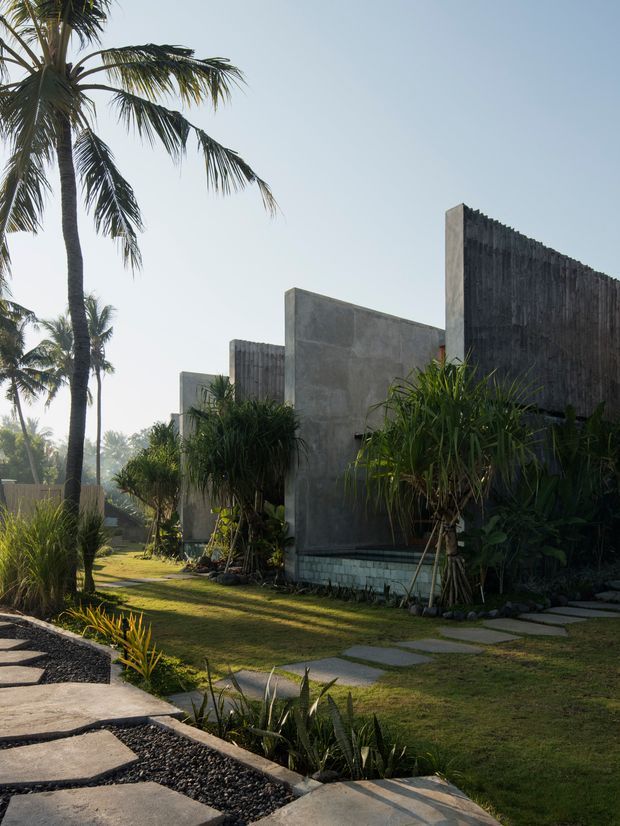 Exterior view of a low-rise modern tropical property with raw concrete walls, tall dark textured wall panels, lush palms and ornamental planting, and a landscaped garden path made from irregular concrete stepping slabs set in dark gravel. The composition shows strong geometric forms softened by dense greenery and warm low-angle sunlight.
