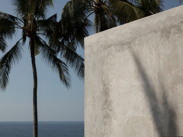 A minimalist coastal exterior detail showing a tall white textured plaster or stucco wall in the foreground, with palm trees silhouetted against a clear sky and the ocean visible in the background. The composition emphasizes the contrast between the organic tropical landscape and the simple architectural plane.