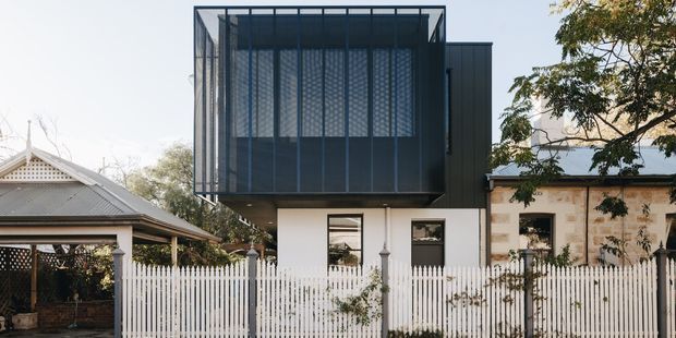 Street-facing exterior view of a contemporary multi-level residential extension, featuring a projecting dark metal upper volume with screened glazing, a light-rendered lower level, and a white picket fence. The building sits among established neighbouring structures and mature trees.