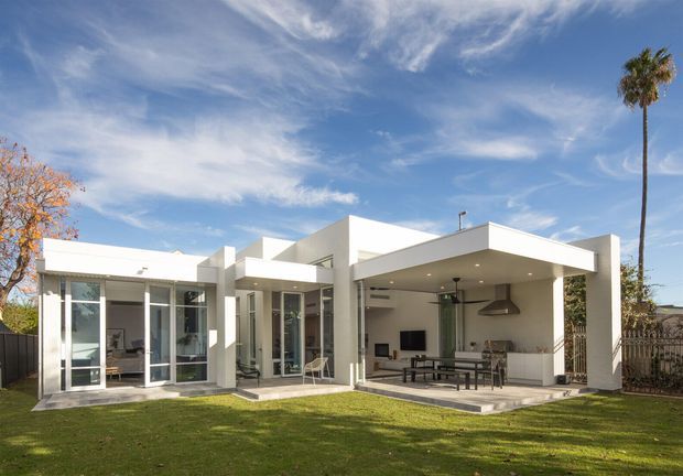 A single-storey modern white-rendered residence viewed from the rear garden. The house has strong geometric volumes, flat roof planes, extensive floor-to-ceiling glazing, and a covered outdoor entertaining area with a dining table, outdoor kitchen, and ceiling fan. A lawn and mature trees surround the home.