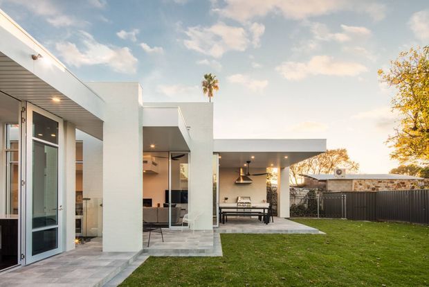 Contemporary single-storey white rendered residence viewed from the garden, featuring crisp cubic forms, flat roofs, extensive glazing, a covered outdoor entertaining area, and a paved terrace. The open carport-like structure includes a dining setting and outdoor fireplace, with a lawn and dark boundary fencing surrounding the home.