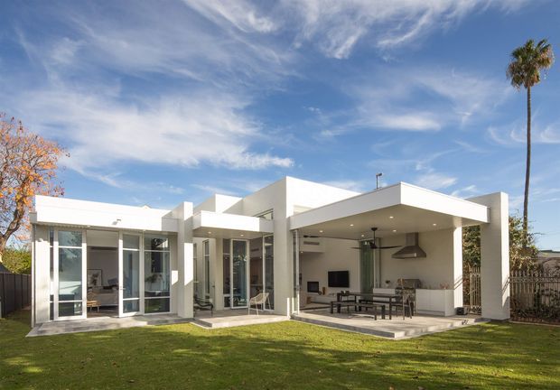 A bright contemporary single-storey home viewed from the rear garden, with crisp white rendered volumes, extensive floor-to-ceiling glazing, a covered outdoor entertaining area, and an integrated outdoor kitchen or barbecue zone. The flat-roofed structure opens onto a broad lawn and includes a shaded dining area with ceiling fan.