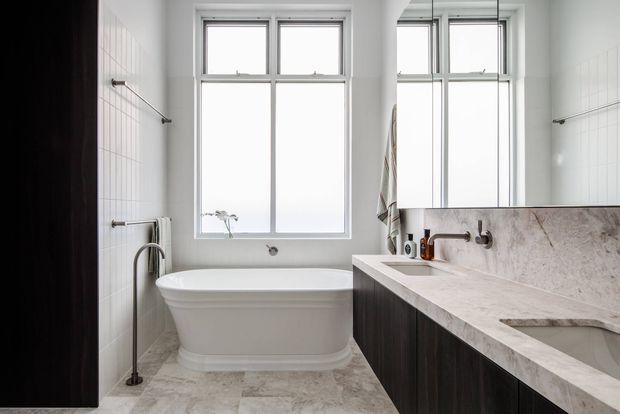Bright, refined bathroom featuring a freestanding white bathtub beneath tall windows, a large dark-wood vanity with pale stone benchtop and integrated basins, pale tiled walls, and a wall-mounted bath tap. A mirror reflects the windows and part of the vanity.