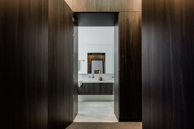 A dark timber-lined passage frames a compact contemporary bathroom. The bathroom features a wall-mounted vanity with a pale stone benchtop, large mirror, white walls, and light stone flooring, viewed through an open doorway.