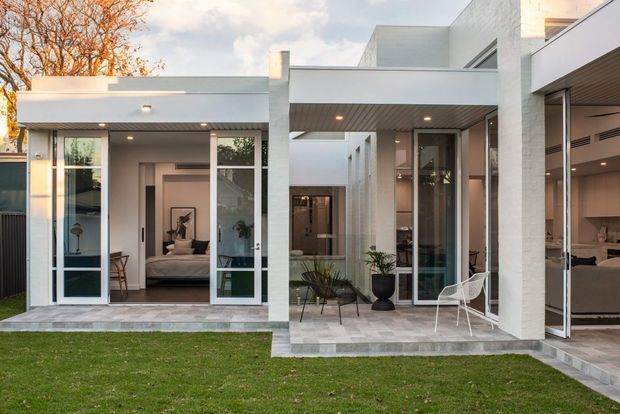 Contemporary white-rendered flat-roof residence viewed from the rear garden, with extensive floor-to-ceiling glazing, covered outdoor terraces, and open bedroom and living areas visible through the glass doors. The design features crisp geometric volumes, recessed lighting, pale tiled paving, and a restrained neutral palette.
