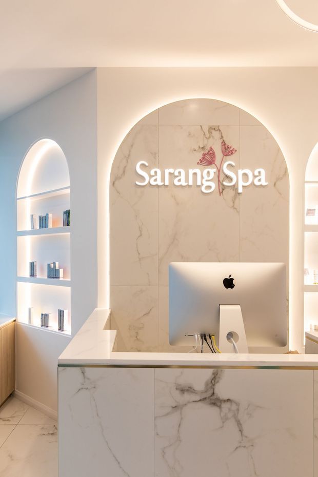 A warmly lit spa reception area featuring a marble-look reception desk and matching arched feature wall with illuminated “Sarang Spa” signage. Built-in arched display niches with books and products flank the desk, while concealed LED lighting creates a soft, refined atmosphere.