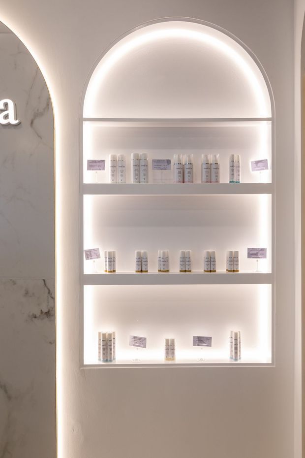 A softly illuminated, arched recessed display niche with three shelves showcasing small product bottles and informational labels. The niche has integrated warm-white LED lighting, white painted surfaces, and a partial marble-look wall panel with curved detailing at the side. The image appears to be from a refined retail, beauty, or wellness interior.