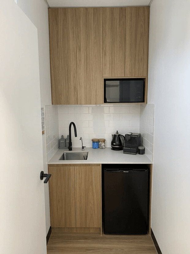 Compact kitchenette fitted into a narrow alcove, with full-height light timber cabinetry, a white subway-tile splashback, pale benchtop, stainless-steel sink, black tapware, microwave, kettle, toaster and under-bench refrigerator. The space is viewed through an open doorway and has a clean, practical contemporary fit-out.