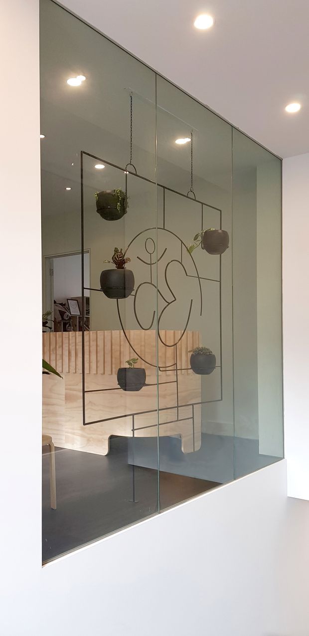 A contemporary interior feature viewed through a large floor-to-ceiling glass partition. The glazing incorporates a thin dark metal geometric and abstract line-art screen, with several suspended dark planters. Behind it is a light timber slatted partition and a dark floor, while recessed ceiling lights illuminate the space. An adjacent room or workspace is partially visible through the glass and opening.