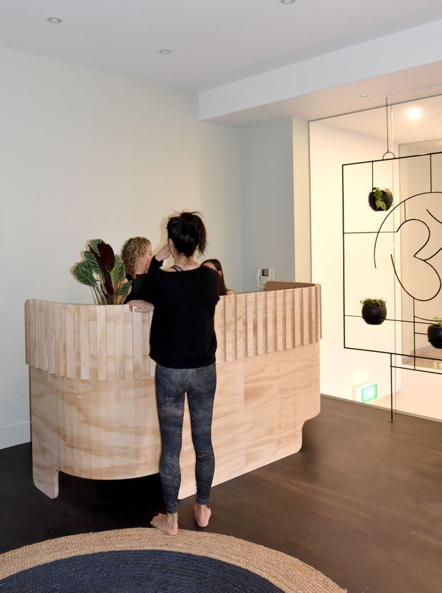 A contemporary reception area with a curved, pale timber reception desk featuring vertical slatted detailing and rounded ends. Several people stand at the desk, which is decorated with leafy plants. The space has white walls and ceiling with recessed downlights, dark timber flooring, and a large black metal geometric room divider with hanging planters and an abstract line motif. A woven circular rug occupies the foreground, and an illuminated exit sign is visible near the floor.
