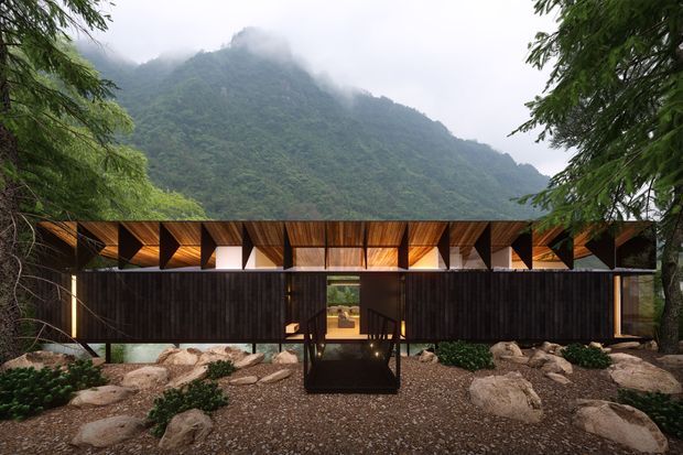 A low, elongated contemporary building set within a lush mountainous landscape. The dark timber-clad facade has extensive glazing and a dramatic folded, geometric timber roof canopy with warm interior lighting visible through the openings. A central entry bridge crosses a rocky landscaped forecourt, with dense tropical vegetation and mist-covered mountains behind.