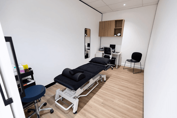 A compact, clean treatment or examination room with a black adjustable treatment table in the foreground, a wheeled stool, a small workstation with computer equipment, wall-mounted timber storage cabinetry, a mirror, and a black visitor chair. The room has white walls, light timber-look flooring, and a suspended ceiling with recessed lighting.