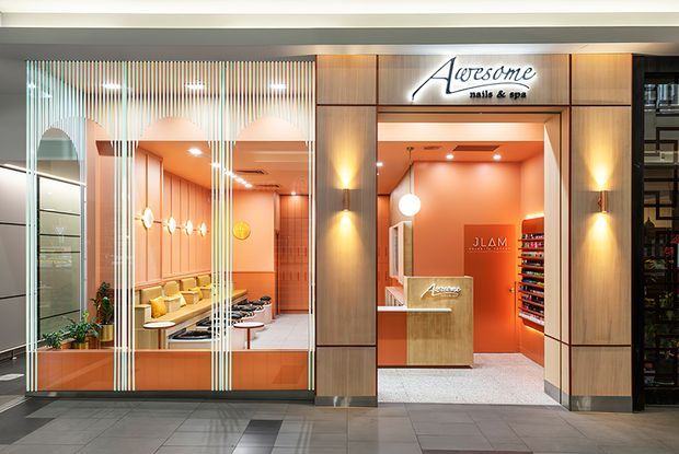 A polished retail salon and spa frontage viewed from a shopping-centre corridor. The open entrance is framed by warm wood-look vertical panels and illuminated signage, with a reception desk and product shelving inside. To the left, full-height glazing with decorative vertical lines reveals peach-toned treatment or service areas, lounge seating, pendant lights, and plants. The space uses warm orange-pink lighting, pale flooring, and a refined contemporary commercial fit-out.