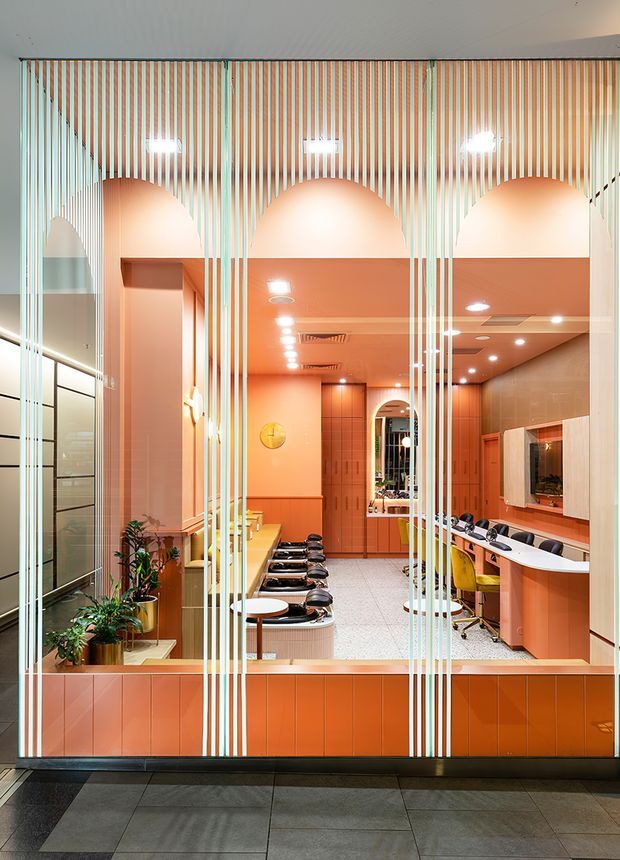A brightly lit commercial interior viewed through a glazed frontage with vertical striped detailing. The space features a coral-orange and peach colour scheme, built-in cabinetry, a long central island with multiple stools, desk workstations, tiled flooring, recessed ceiling lighting, and a rear service or display area. The composition and fixtures suggest a salon, beauty studio, or similar retail service environment.