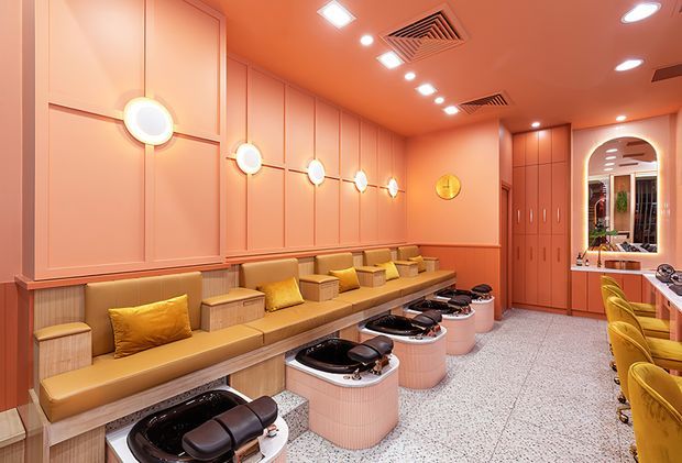 A warmly lit, highly stylised salon interior with a long built-in mustard upholstered bench along a peach and coral paneled wall. Several rounded pale pink manicure or treatment stations with black basins occupy the centre, while matching yellow salon chairs and counters line the right side. Circular wall sconces, recessed lighting, terrazzo-style flooring, extensive built-in cabinetry, and an arched opening contribute to a playful retro-luxe commercial design.