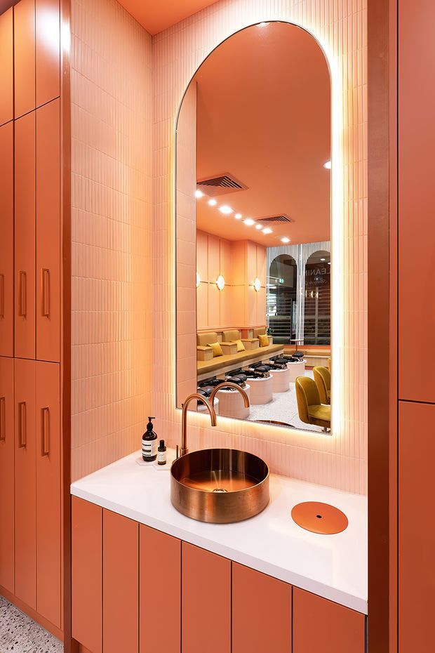 A highly stylised powder-room vanity in warm coral and terracotta tones. Full-height narrow rectangular wall tiles surround a large illuminated arched mirror, with matching built-in cabinetry framing the space. The white vanity benchtop holds a round copper vessel basin, curved copper tapware and a circular orange drain or accessory. The mirror reflects a richly coloured commercial-looking interior with banquette seating, pendant or wall lights, and additional basins, suggesting a hospitality venue or public washroom.